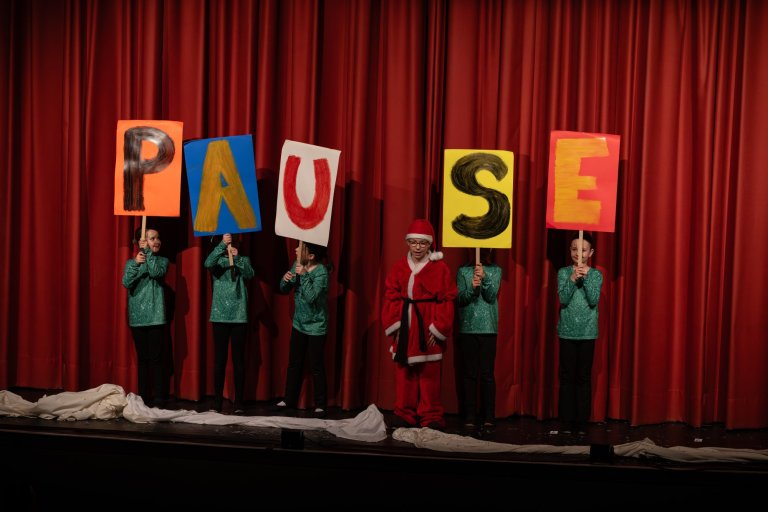 Kinder in grünen Kostümen halten bunte Buchstabentafeln "PAUSE" vor rotem Theatervorhang, Nikolaus in Kostüm in der Mitte.