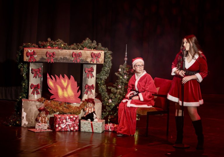 Zwei Darsteller in roter Weihnachtskleidung auf Bühne mit dekoriertem Kamin und Geschenken.