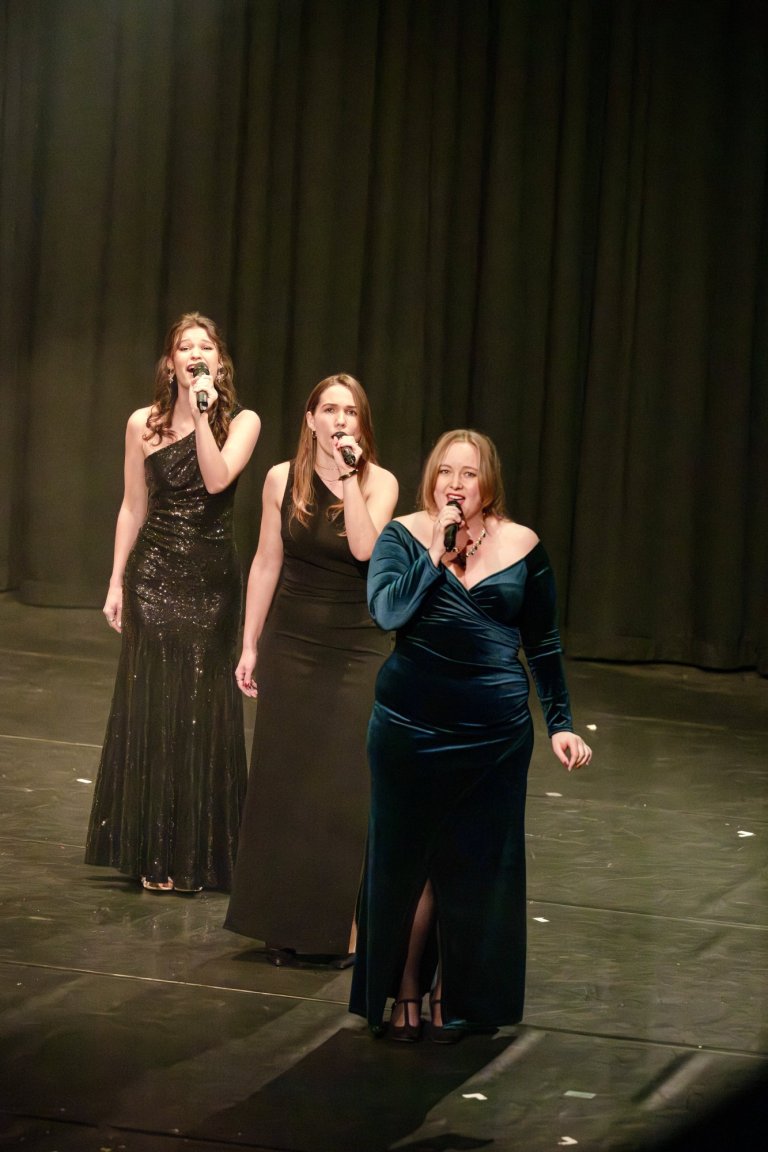 Drei Sängerinnen in eleganten Abendkleidern performen auf der Bühne mit Mikrofonen vor dunklem Vorhang.