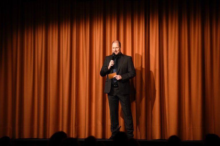 Hauke Wendt in schwarzem Anzug mit Mikrofon auf Bühne vor orangefarbenem Vorhang präsentiert Musicalschule-Event.