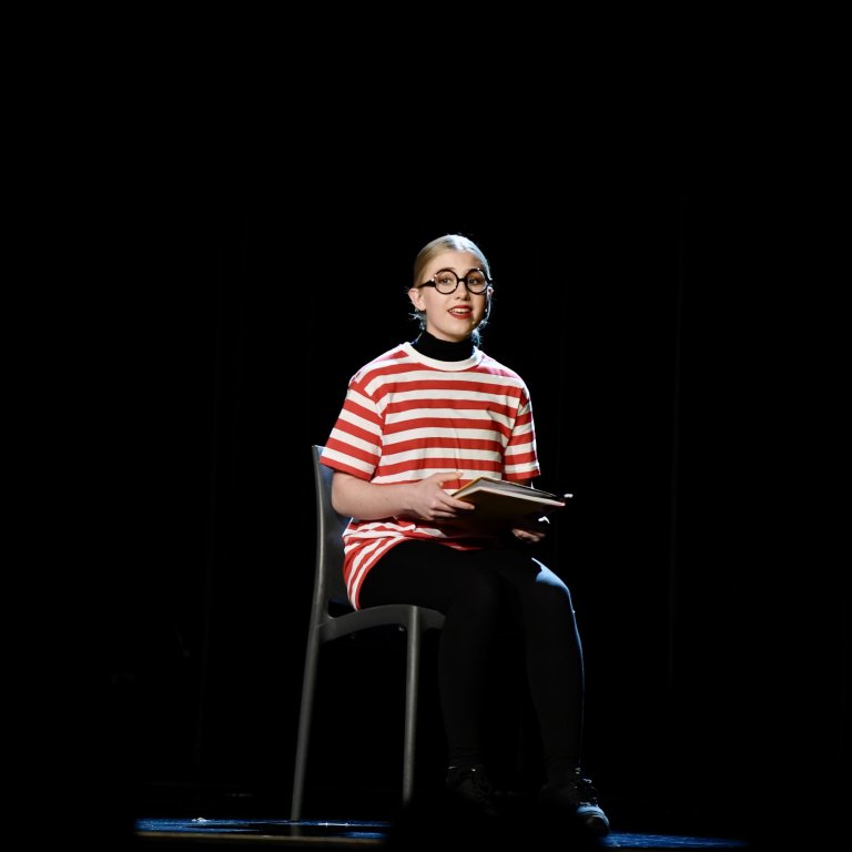 Junge Person in rot-weiß gestreiftem Shirt mit Brille sitzt auf Stuhl und hält aufgeschlagenes Buch während Musiktheater-Aufführung.