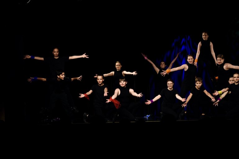 Gruppe junger Tänzer in schwarzen Outfits mit bunten Armbändern auf der Bühne in synchroner Choreographie.