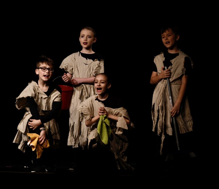 Fünf Kinder in zerfetzter Kostümierung singen gemeinsam auf einer dunklen Bühne während einer Musicalaufführung.