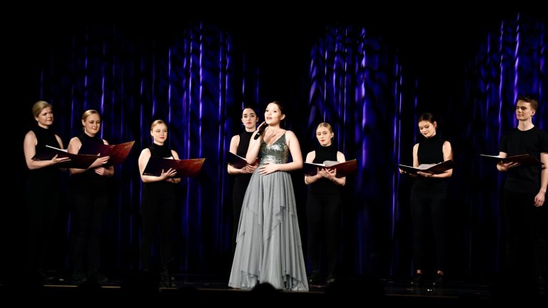 Musicalschule Ahrensburg: Sänger und Sängerinnen in schwarzen Outfits mit Noten-Ordnern präsentieren Chorperformance auf Bühne.