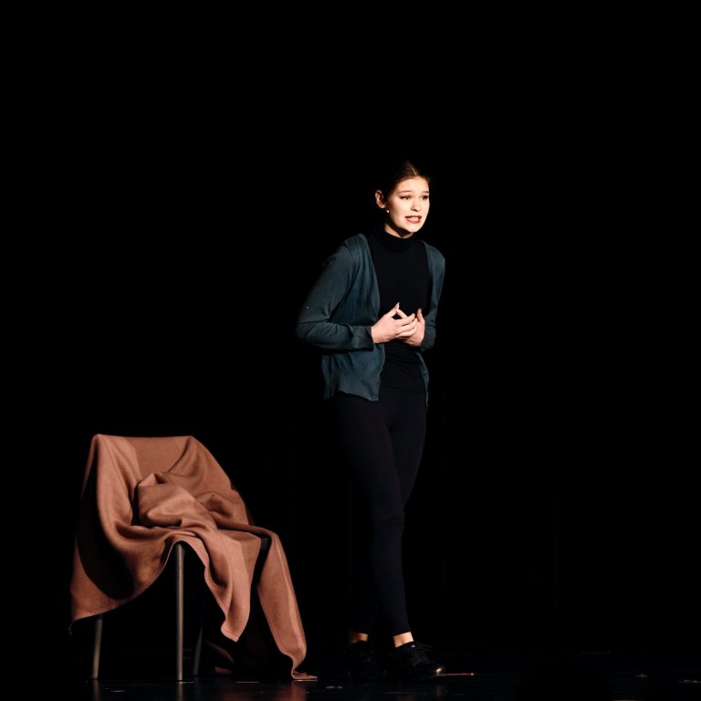Junge Schauspielerin auf schwarzer Bühne neben braunem Stuhl während Musicalaufführung steht.