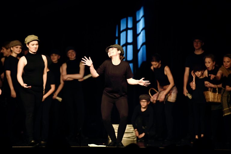 Jugendliche Darsteller in schwarzer Kleidung und Mützen auf Theaterbühne mit bleuchteter Fensterkulisse während Musicalaufführung.