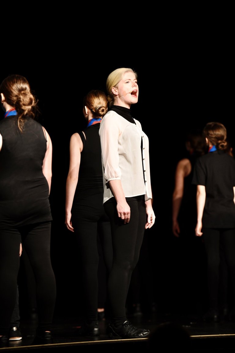 Schüler der Musicalschule Ahrensburg treten auf dunkler Bühne auf, singen gemeinsam im Chor während Musiktheater-Aufführung.