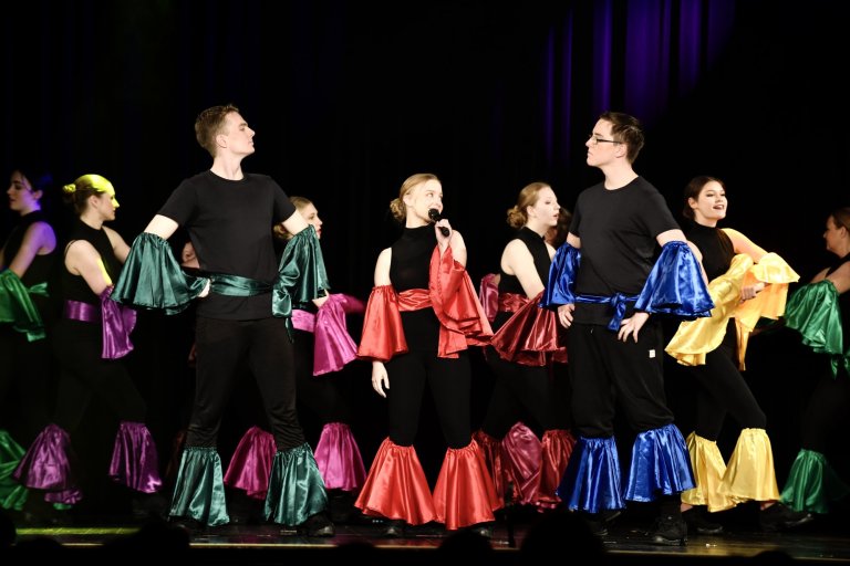 Musicalschüler in schwarzer Kleidung mit farbigen Seidencapes performen gemeinsam auf der Bühne beim Musical "All Together Now".