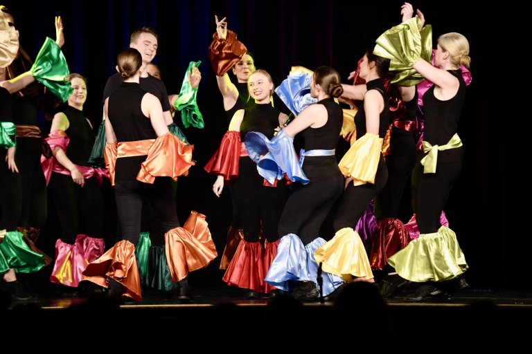 Kindertheatergruppe führt "All Together Now" auf: Kinder in schwarzen Outfits mit bunten Seidenschals tanzen synchron auf Bühne.