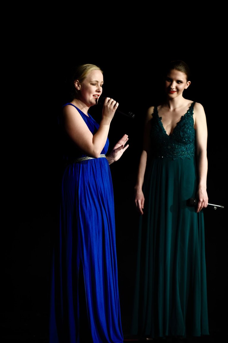 Zwei Sängerinnen in eleganten Abendkleidern performen gemeinsam auf der Bühne während eines Musicalschul-Auftritts.