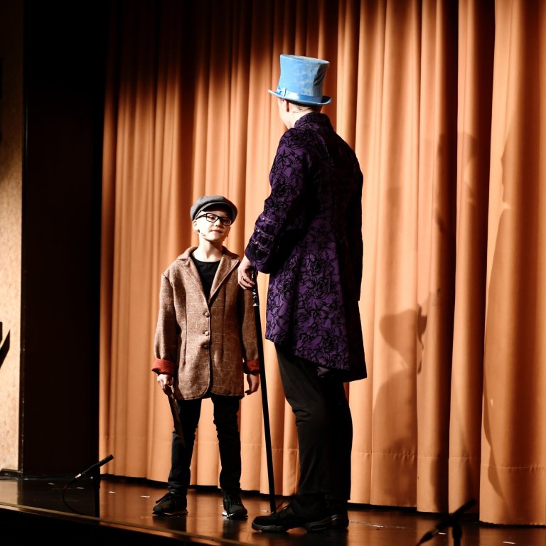 Zwei Performer auf Theaterbühne: Kind in braunem Blazer und Erwachsener mit blauem Zylinder vor goldenem Vorhang.