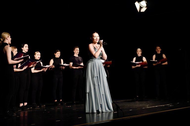 Solistin in grauem Kleid singt mit Mikrofon, begleitet von Chor in schwarzer Kleidung auf Theaterbühne.