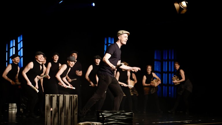 Tanzgruppe in schwarzen Outfits führt choreografierte Musical-Szene auf Bühne mit blauer Beleuchtung auf.