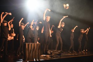Gruppe von Schauspielern in schwarzer Kleidung und Hüten auf Bühne mit dramatischer Beleuchtung während Musicalaufführung.