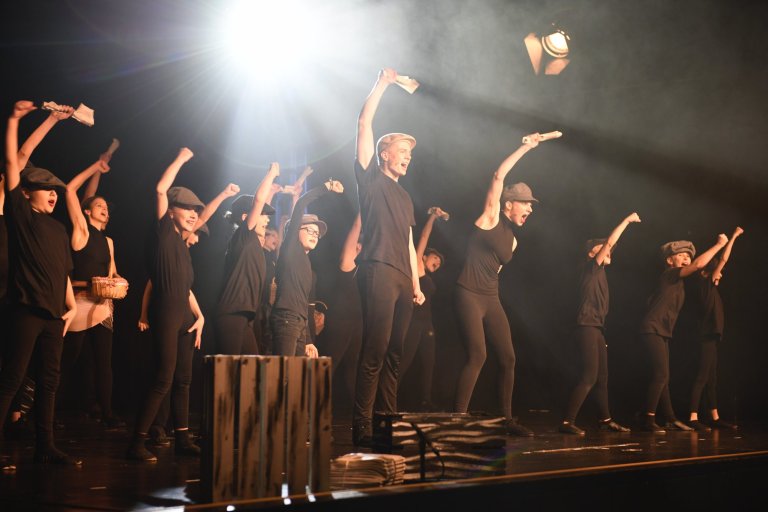 Gruppe von Schauspielern in schwarzer Kleidung und Hüten auf Bühne mit dramatischer Beleuchtung während Musicalaufführung.