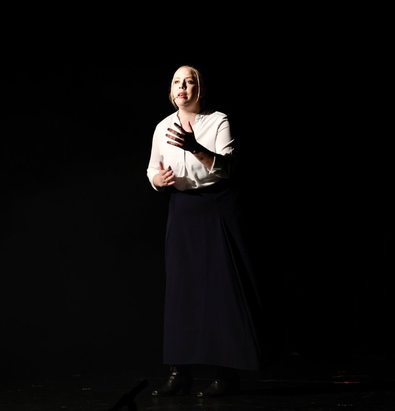 Schauspielerin auf dunkler Bühne in weißer Bluse und schwarzem Rock performt emotional mit ausdrucksstarker Gestik.