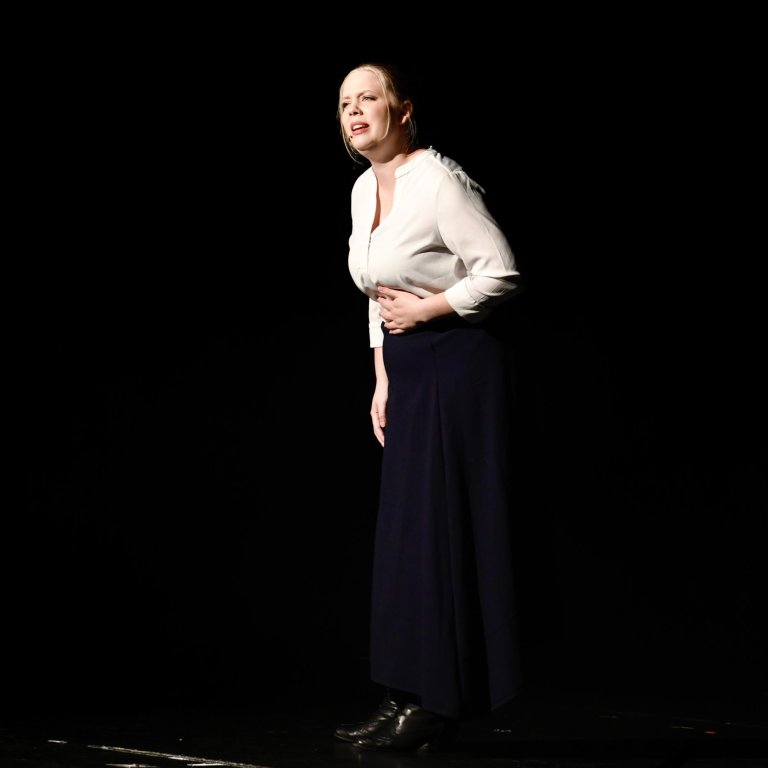Frau in weißer Bluse und dunkler Hose steht auf Bühne, performt beim Musical All Together Now.