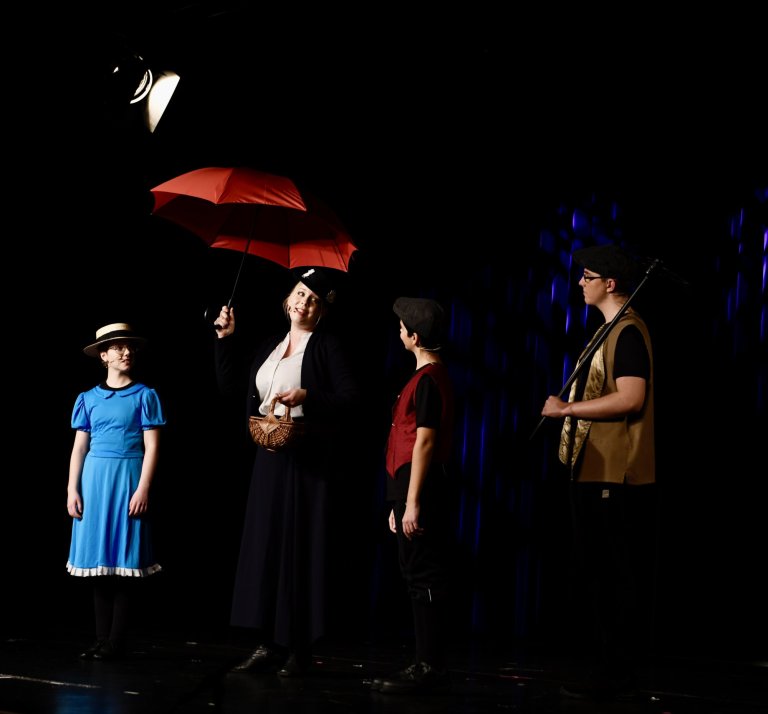 Theaterszene mit vier Schauspielern auf dunkler Bühne, einer hält roten Regenschirm, musikalische Inszenierung.
