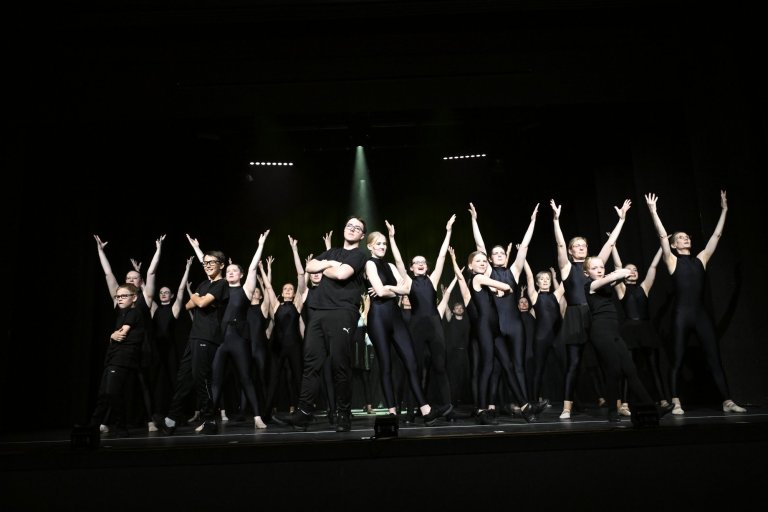 Gruppe von Schülern in schwarzer Kleidung auf Bühne mit erhobenen Armen, Jubiläumsshow MSA10 Musicalschule Ahrensburg.