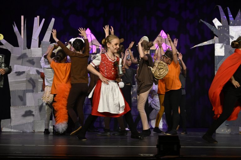 Kindermusicalgruppe auf Bühne mit roten und orangefarbenen Kostümen, Bühnendeko mit Häusern, Jubiläumsshow MSA10.