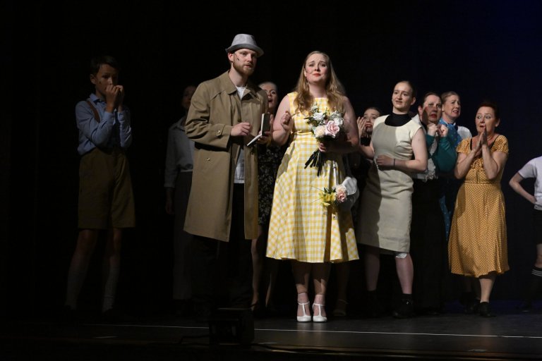 Musicalschule-Ensemble auf Bühne: Frau im gelben Kleid mit Blumenstrauß, Mann in Trenchcoat und Hut, weitere Darsteller im Hintergrund.