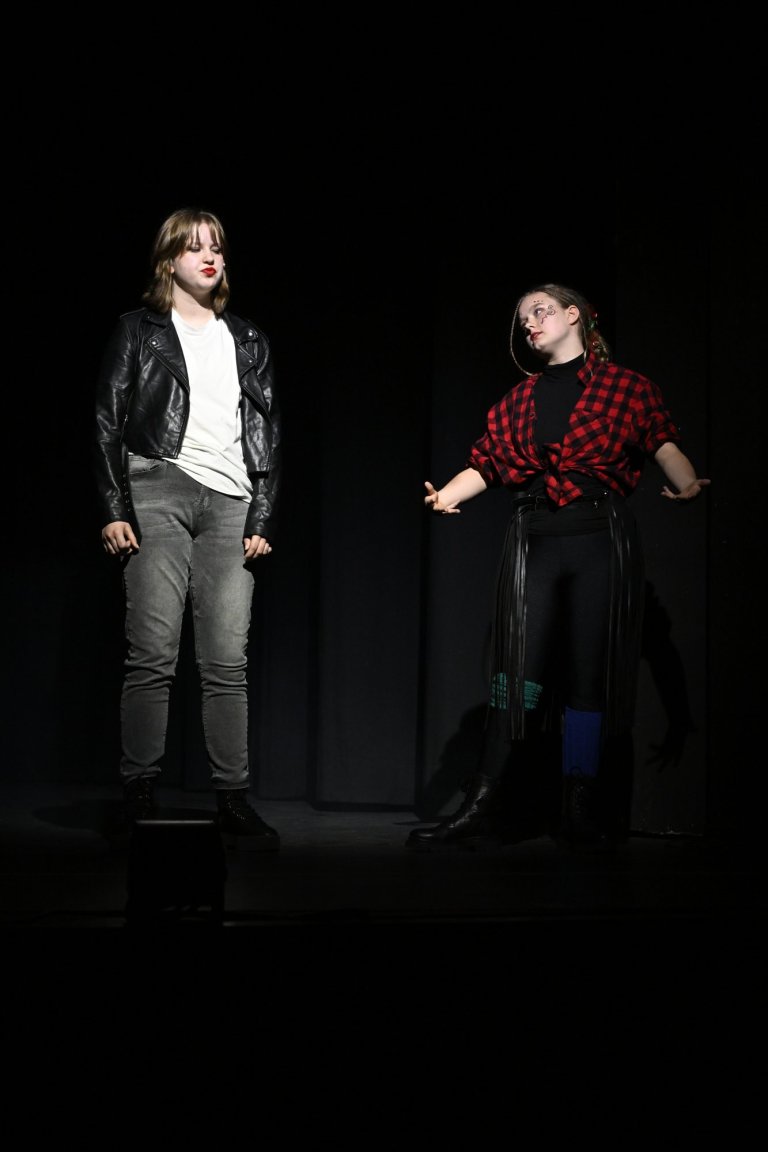 Zwei Performer auf dunkler Bühne: Frau in schwarzer Lederjacke und grauer Hose, Mann in rotem Karohemd, dramatische Theaterszene.