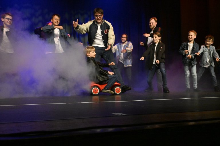 Gruppe von Kindern und Erwachsenen auf Bühnebühne mit Nebeleffekt, Kind auf rotem Rutschauto, Jubiläumsshow MSA10.