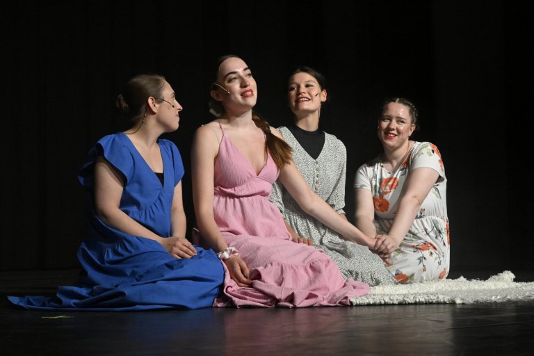 Vier junge Darstellerinnen sitzen auf der Bühne in farbigen Kleidern und schauen nach oben, Musicalschule Ahrensburg.