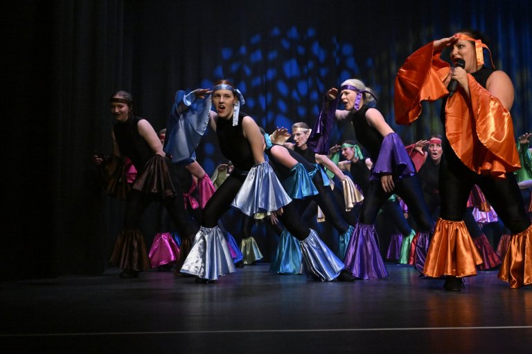 Musicalschüler auf Bühne in farbigen Kostümen singen während Jubiläumsshow MSA10 mit bunten Röcken und Caps.