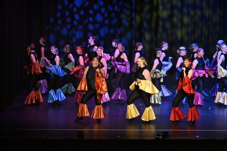 Große Gruppe Tänzer auf Bühne in bunten Kostümen mit glänzenden Umhängen, Jubiläumsshow Musicalschule Ahrensburg.