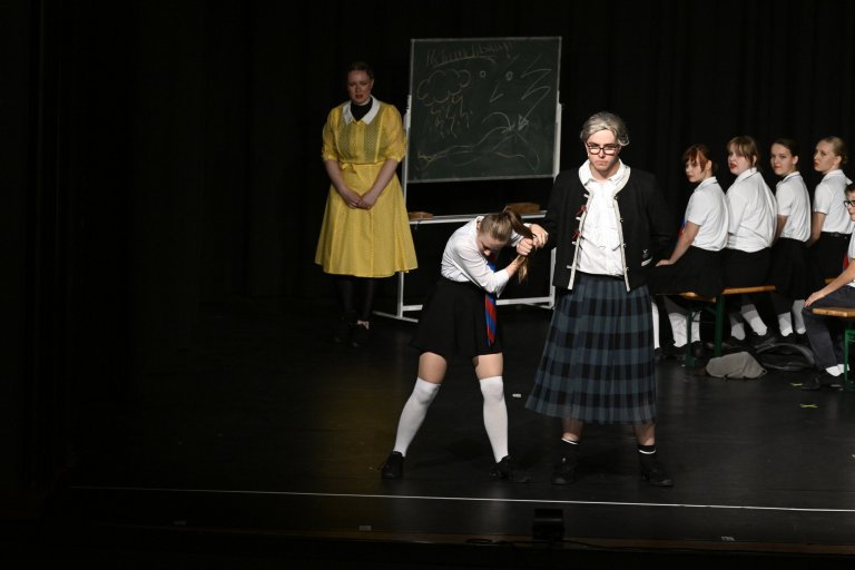 Schulmusical-Aufführung: Lehrerin und Schülerinnen in Schuluniform auf Bühne mit Tafel, Jubiläumsshow MSA10.