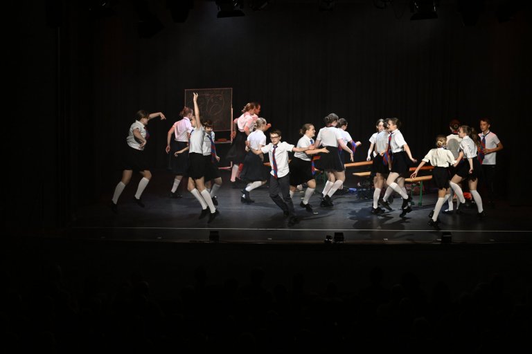 Kinderensemble der Musicalschule Ahrensburg tanzt energiegeladen auf Bühne, Jubiläumsshow MSA10 mit Schuluniformen und Requisiten.