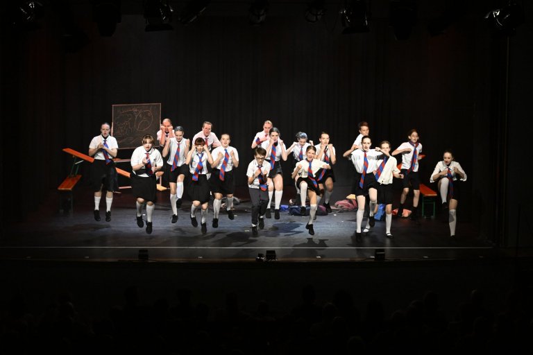 Gruppe junger Performer springt auf Bühne, trägt weiße Hemden mit roten-blauen Medaillen, Jubiläumsshow der Musicalschule Ahrensburg.