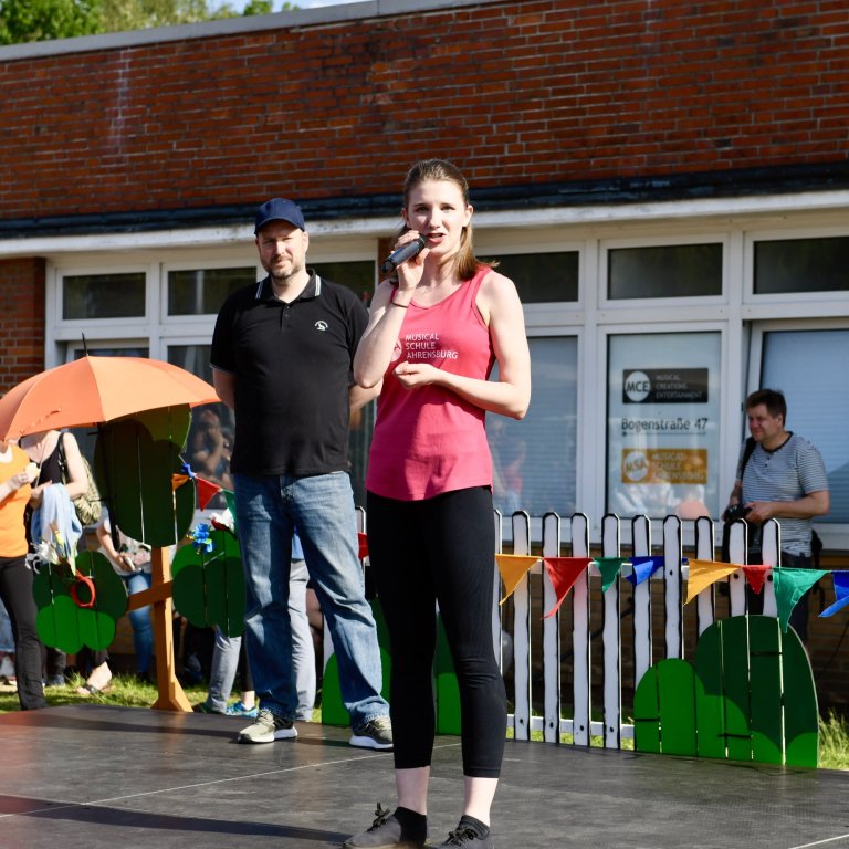 Moderatorin mit Mikrofon beim MSA Sommerfest 2022 auf der Bühne vor Schulgebäude und Publikum.
