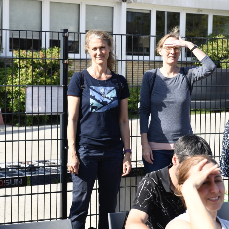 Vier Personen stehen vor einem schwarzen Zaun vor einer modernen Schule mit weißen Fenstern.