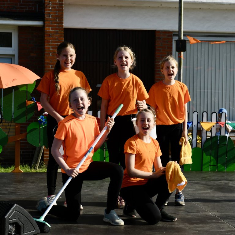 Sechs Kinder in orangenen T-Shirts beim Sommerfest der Musicalschule Ahrensburg, fröhlich posierend mit Requisiten.
