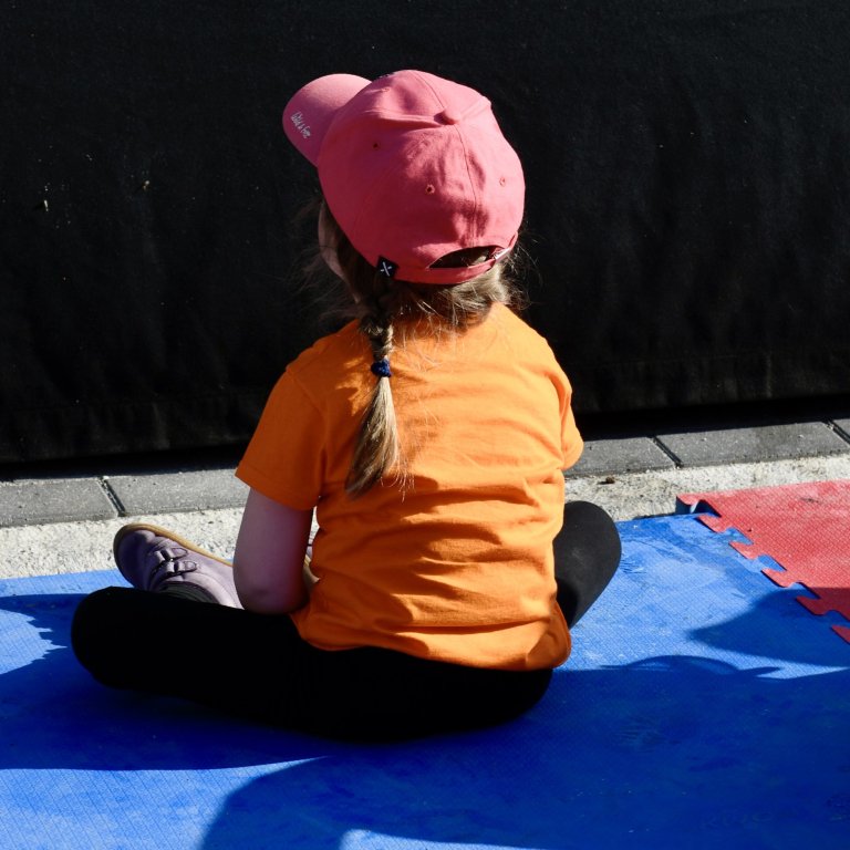 Kind sitzt in oranger Kleidung und rosa Kappe auf blauer Matte beim MSA Sommerfest 2022.
