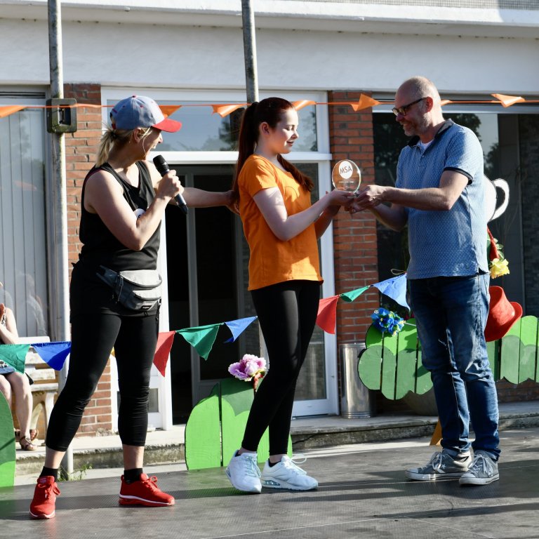 Preisverleihung beim MSA Sommerfest 2022: Frau überreicht Preis an Gewinnerin in orangem Shirt, Moderatorin mit Mikrofon.