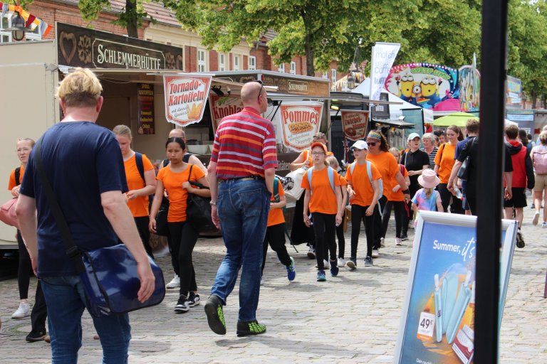 Kinder in orangefarbenen Shirts laufen beim Lindenfest Ludwigslust, umgeben von Marktständen und Besuchern.