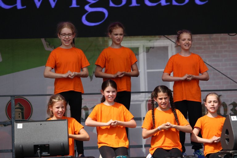 Gruppe junger Schüler in orangen Shirts beim Lindenfest Ludwigslust, Musicalschule Ahrensburg Auftritt auf Bühne.