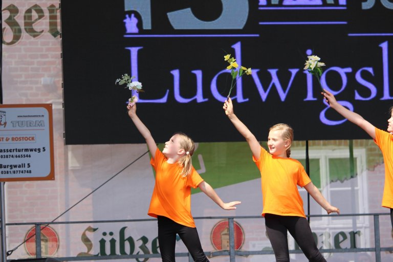 Junge Tänzerinnen in orange Shirts halten Blumensträuße beim Lindenfest Ludwigslust auf der Bühne.
