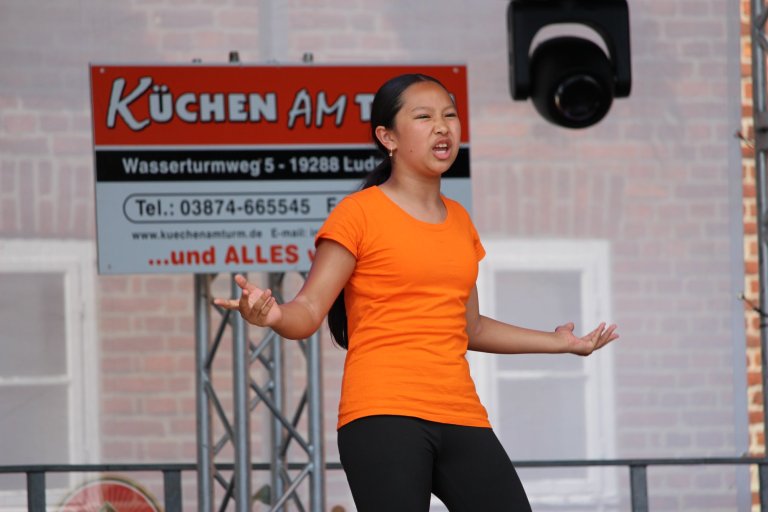Junge Performerin in orangefarbenem Shirt präsentiert energiegeladene Tanzperformance auf Bühne beim Lindenfest Ludwigslust.