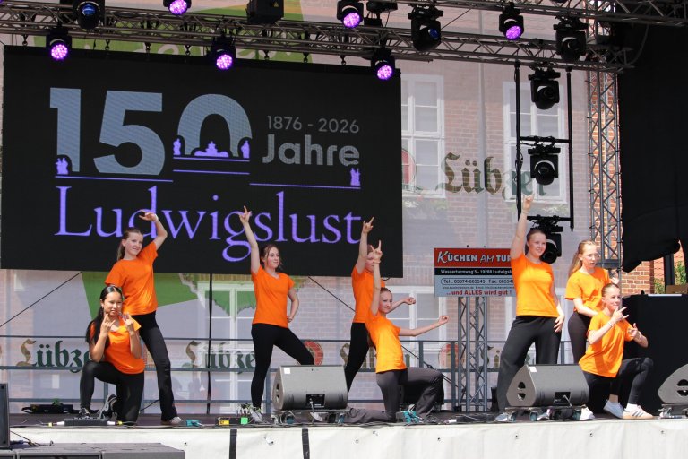 Kinder in orangenen Shirts tanzen auf Bühne zum Ludwigslust 150-Jahre-Jubiläum auf dem Lindenfest.
