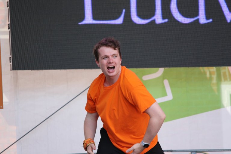 Junger Mann in orangem Shirt tanzt begeistert beim Lindenfest Ludwigslust auf der Bühne der Musicalschule Ahrensburg.