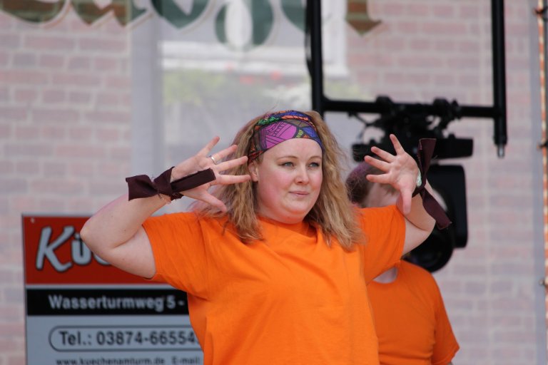 Mädchen in orangem Shirt mit buntem Kopftuch performt beim Lindenfest Ludwigslust der Musicalschule Ahrensburg.