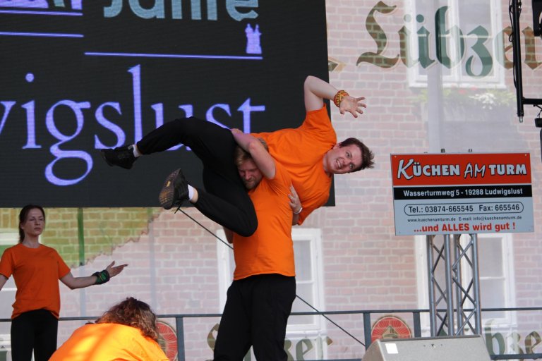 Akrobatischer Auftritt bei Lindenfest Ludwigslust: Performer in orangem Shirt hebt Kollegen hochakrobatisch auf Bühne.