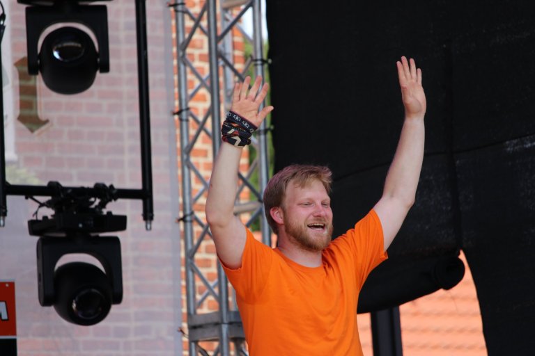 Mann in orangem Shirt mit erhobenen Armen auf Bühne beim Lindenfest Ludwigslust, Bühnentechnik im Hintergrund.