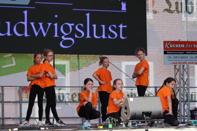 Gruppe junger Performer in orangefarbenen Shirts auf Bühne während Lindenfest Ludwigslust Musikaufführung.