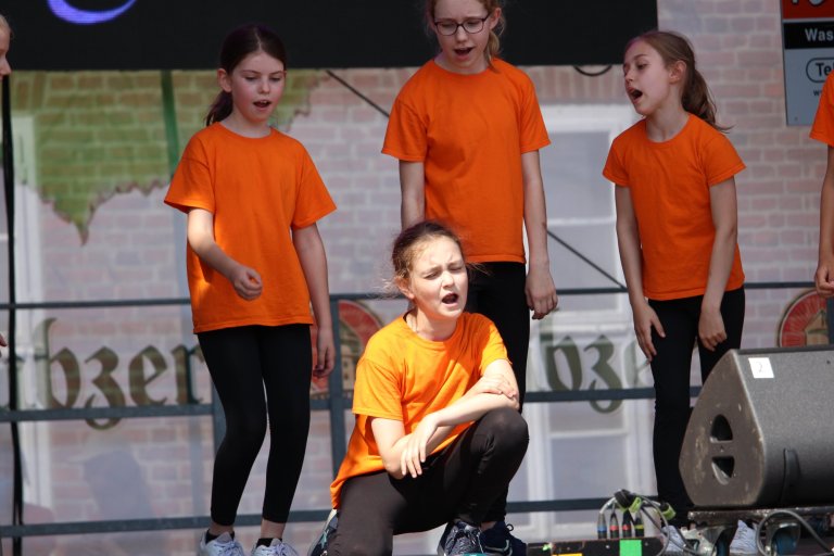 Kindermusicalgruppe in orangenen Shirts performt auf Bühne beim Lindenfest Ludwigslust der Musicalschule Ahrensburg.