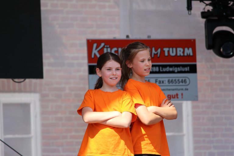 Zwei Mädchen in orangenen T-Shirts tanzen beim Lindenfest Ludwigslust vor Kulisse der Musicalschule Ahrensburg.